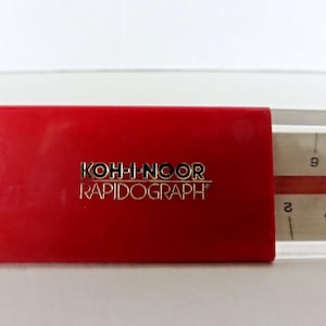 May include: A close-up of a red Koh-I-Noor Rapidograph pen case. The brand name is in gold lettering. A portion of a ruler is visible on the right side of the image, with the number 2 and 9 in view.