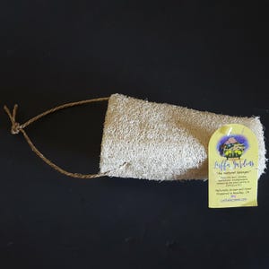 May include: A natural loofah sponge with a woven texture, attached to a brown string loop. A yellow tag with the text "Luffa Gardens" and "Au Naturel Sponges" is attached to the sponge. The tag also includes the text "Naturally Grown and Hand Prepared in Reedley, CA".