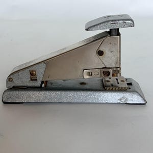 May include: A vintage silver stapler with a rectangular base and a curved top. The stapler has a lever for pressing staples and a textured finish. The design is classic and functional, suitable for office or home use.