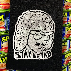 Könnte beinhalten: Ein schwarzer Patch mit einer weißen Illustration einer Person mit einer großen Afro-Frisur, die eine Brille und einen Schnurrbart trägt. Der Text "STAY WEIRD" ist unter der Illustration gedruckt.
