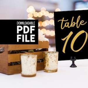 May include: Black table number card with gold text that reads "table 10". The card is sitting on a black stand and is on a white table with a wooden crate, white flowers, and two gold candle holders.