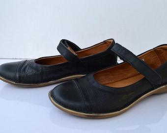 May include: A pair of black leather flats with a strap across the top. The shoes have a brown sole and a perforated design.