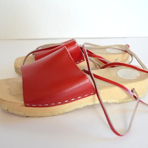 May include: A pair of red leather sandals with wooden soles. The sandals have a wide red strap across the top and thin red straps that wrap around the ankle. The wooden soles are light-colored and have a slight platform.