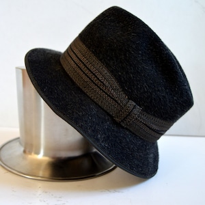 Op de afbeelding: Een zwarte fedora hoed met een bruin en zwart gestreept lint. De hoed ligt op een zilveren metalen voet.