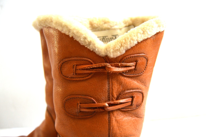 Puede incluir: Una bota de cuero color canela con un forro de piel de oveja color crema en la abertura superior. El exterior de la bota est&aacute; asegurado con dos cierres de palanca horizontales de cuero, cada uno con un lazo y una barra de cuero a juego.