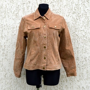 May include: A tan suede jacket featuring a pointed collar, long sleeves, and a front button closure. The garment includes two chest pockets with buttoned flaps and metallic buttons throughout, displayed on a black mannequin torso.