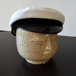 Op de afbeelding: Een witte matrozenhoed met een zwarte rand en band rust op een bespikkelde keramische vaas in de vorm van een hoofd. De hoed heeft een afgeronde kroon en een glanzend, zwart vizier. De vaas heeft een gezicht gebeeldhouwd aan de voorkant.