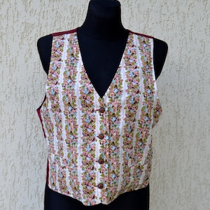 Op de afbeelding: Een vest met bloemenprint met een V-hals en houten knopen. Het vest heeft een patroon van roze, blauwe en groene bloemen op een witte achtergrond met verticale strepen. Het vest heeft een bordeauxrode voering en wordt gepresenteerd op een zwarte mannequin.