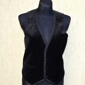 May include: A black velvet waistcoat with a notched lapel and a five-button closure. The waistcoat has two front pockets and a satin trim. It is displayed on a black mannequin.