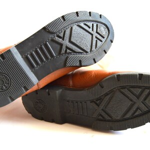 Puede incluir: Un par de botas de cuero color canela con suelas de goma negra. Las suelas presentan un patr&oacute;n de rodadura profundo con un dise&ntilde;o de trama cruzada en el centro y un logotipo circular en el tal&oacute;n que dice "JAM & SHORT" con una estrella.