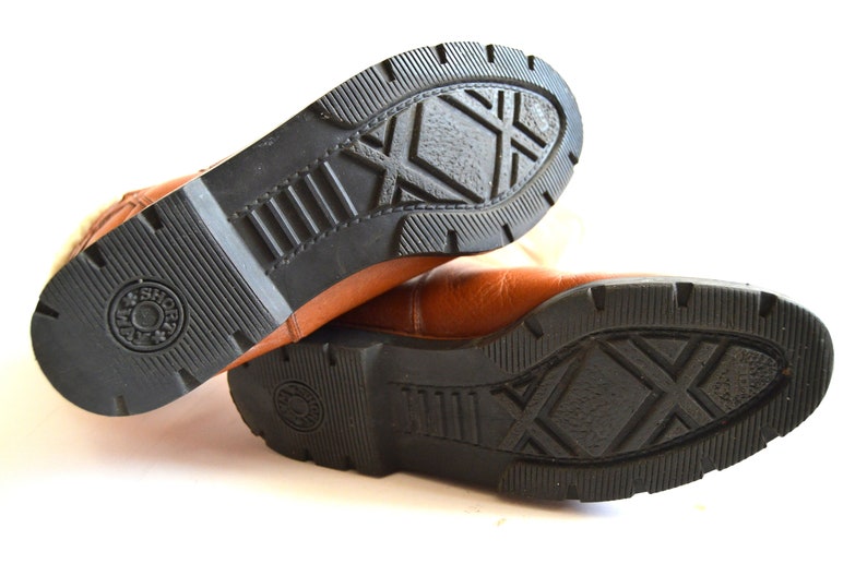 Puede incluir: Un par de botas de cuero color canela con suelas de goma negra. Las suelas presentan un patr&oacute;n de rodadura profundo con un dise&ntilde;o de trama cruzada en el centro y un logotipo circular en el tal&oacute;n que dice "JAM & SHORT" con una estrella.