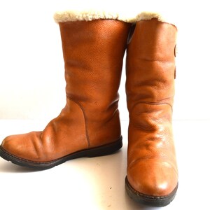 Puede incluir: Un par de botas de media ca&ntilde;a en color marr&oacute;n claro, con un exterior de cuero texturizado, punta redondeada y una suela gruesa de goma marr&oacute;n oscuro. La abertura superior de cada bota est&aacute; forrada con un suave material de felpa color crema.