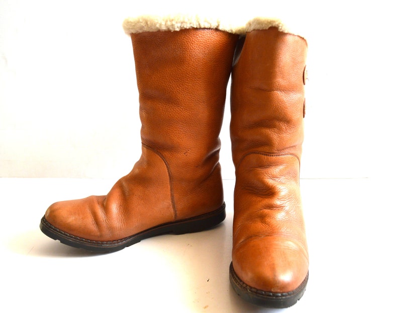 Puede incluir: Un par de botas de media ca&ntilde;a en color marr&oacute;n claro, con un exterior de cuero texturizado, punta redondeada y una suela gruesa de goma marr&oacute;n oscuro. La abertura superior de cada bota est&aacute; forrada con un suave material de felpa color crema.
