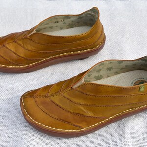 May include: A pair of brown leather slip-on shoes. The shoes have a rounded toe and a stitched design across the top. The soles are a darker brown, and the stitching is white. The interior has a patterned lining.