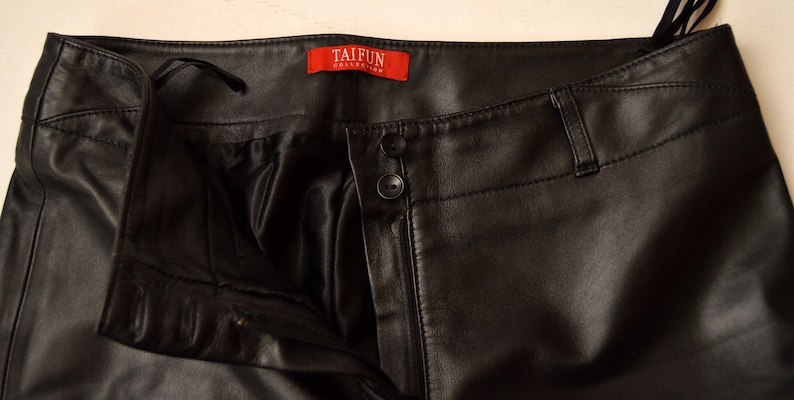 May include: Black leather pants with a zipper fly and button closure. The pants have a label that reads "TAIFUN COLLECTION".