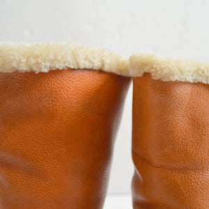 Puede incluir: Un par de botas de cuero color canela con una superficie texturizada y un forro de piel sint&eacute;tica color crema en la abertura superior. La bota derecha muestra dos cierres de palanca de cuero verticales en el lateral.