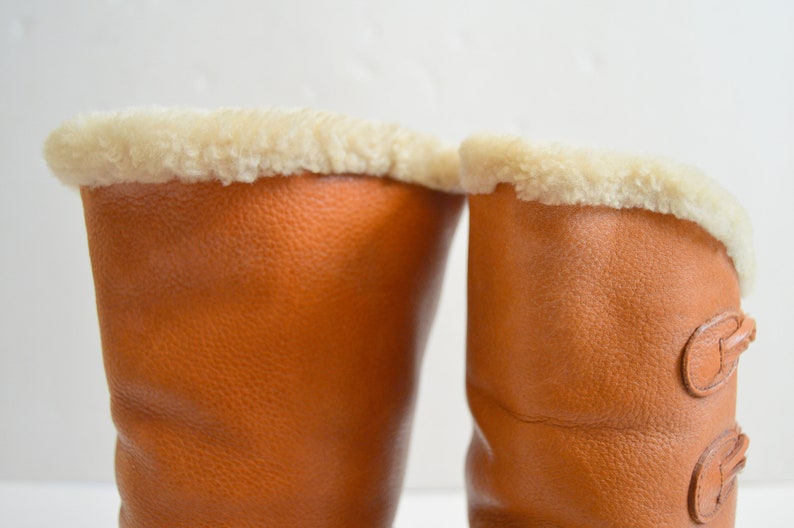 Puede incluir: Un par de botas de cuero color canela con una superficie texturizada y un forro de piel sint&eacute;tica color crema en la abertura superior. La bota derecha muestra dos cierres de palanca de cuero verticales en el lateral.