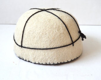 May include: A cream-colored, dome-shaped hat with black stitching and black lines forming a grid pattern across the top. The hat has a small black tassel on the side. The hat appears to be made of a soft, textured material.