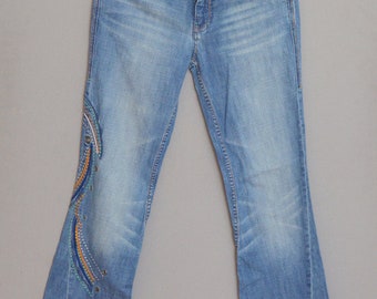 May include: A pair of blue denim jeans with a colorful embroidered design on the left leg. The jeans are hanging on a gold hanger against a gray background.