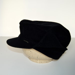 May include: A black hat made from a wool-like material, featuring a short brim and a small, rectangular metal detail. The hat is placed on a speckled, off-white ceramic bowl. The design includes a structured crown and a slightly angled brim.