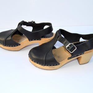 Pode incluir: Um par de tamancos de couro preto com design de tira em T. Os tamancos têm solas de madeira e saltos pequenos. O couro é costurado na base de madeira com linha branca. Os tamancos estão em um fundo branco.