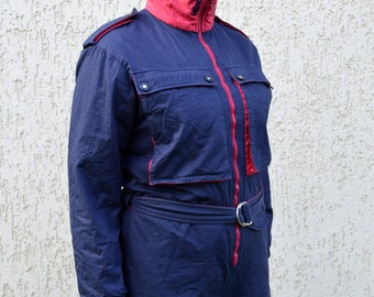 May include: A vintage navy blue ski suit with red accents. The one-piece suit features a high collar, a front zipper, and a belt with a silver buckle. The suit has two chest pockets and red trim along the zipper and pockets.