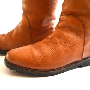 Puede incluir: Un par de botas de media ca&ntilde;a confeccionadas en cuero color canela con una textura granulada visible. Las botas presentan punteras redondeadas, detalles de paneles cosidos en los laterales y suelas gruesas de goma dentada marr&oacute;n oscuro.