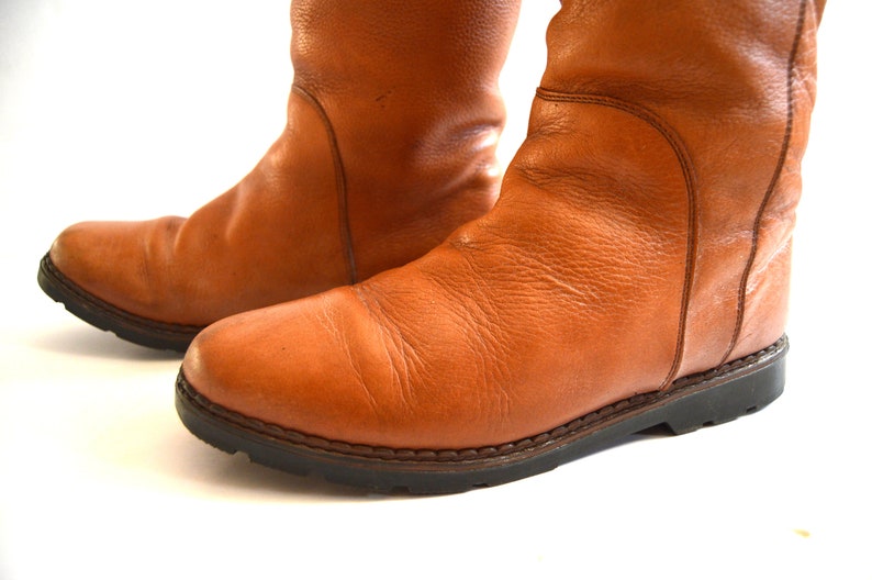 Puede incluir: Un par de botas de media ca&ntilde;a confeccionadas en cuero color canela con una textura granulada visible. Las botas presentan punteras redondeadas, detalles de paneles cosidos en los laterales y suelas gruesas de goma dentada marr&oacute;n oscuro.