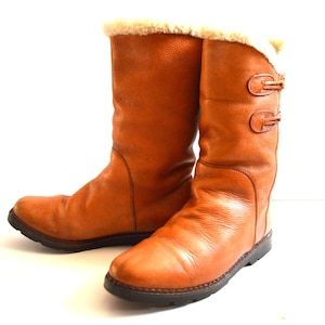 Puede incluir: Un par de botas de media ca&ntilde;a hechas de cuero color canela, con un forro de piel sint&eacute;tica color crema en la abertura superior. Cada bota presenta dos cierres de palanca decorativos en el lado exterior y una suela de goma marr&oacute;n oscuro.