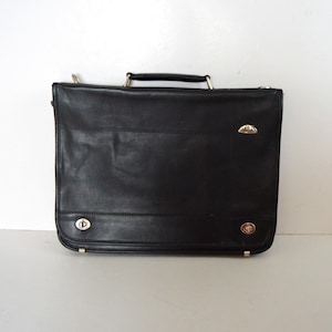 Puede incluir: Maletín de cuero negro con asa superior y herrajes plateados. La bolsa rectangular presenta un cierre de solapa y detalles decorativos plateados. Un accesorio de negocios clásico.