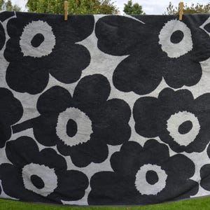 May include: A large beach towel with a striking floral design. The towel displays large, stylized black flowers with white centers on a grey background. It is hanging on a washing line, demonstrating its size and pattern. The edges have a dark grey border.