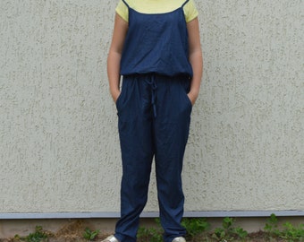 May include: A dark blue jumpsuit with a drawstring waist and pockets. The jumpsuit is worn over a yellow t-shirt. The model is wearing silver sandals. The outfit is casual and comfortable.
