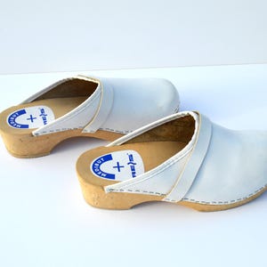 May include: A pair of white leather clogs with wooden soles. The clogs have a rounded toe and a strap across the top. The soles are light brown, and the clogs have a label with text and a plus sign.