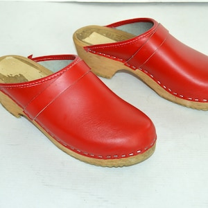 May include: A pair of red leather clogs with wooden soles. The clogs have a classic design with a rounded toe and a strap across the top. The soles are made of light-coloured wood and are approximately 5 cm high.