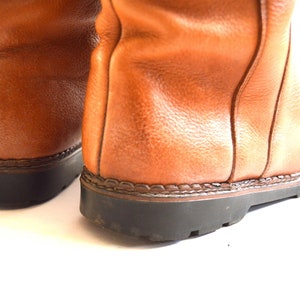Puede incluir: Un par de botas de cuero color canela con una superficie texturizada, costuras verticales visibles a lo largo del tal&oacute;n trasero y una suela de goma negra gruesa con un detalle de ribete cosido que une el cuero superior a la base.