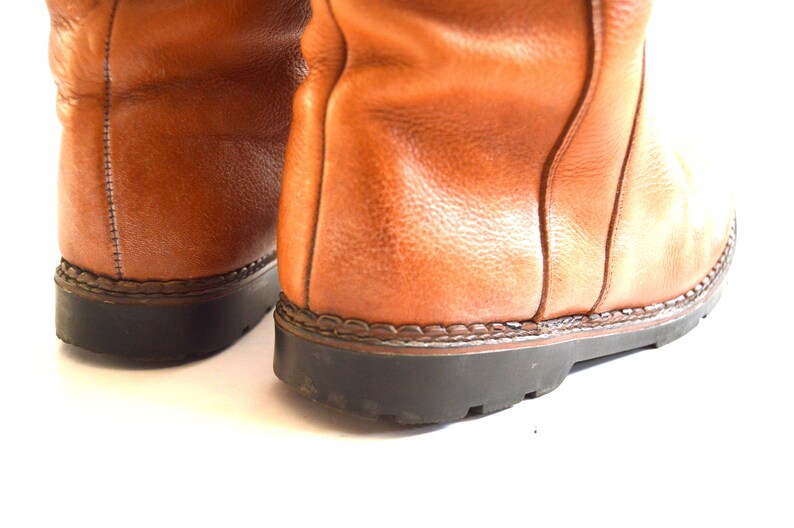 Puede incluir: Un par de botas de cuero color canela con una superficie texturizada, costuras verticales visibles a lo largo del tal&oacute;n trasero y una suela de goma negra gruesa con un detalle de ribete cosido que une el cuero superior a la base.