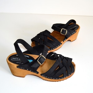 May include: Black leather sandals with a woven design and a wooden platform sole. The sandals have a buckle closure and are made in Sweden. The brand name "MOHE" is visible on the sole.