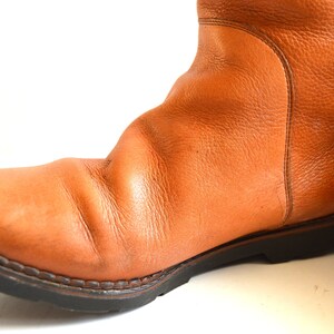 Puede incluir: Una bota de cuero color canela con superficie texturizada, punta redondeada y una suela gruesa de goma negra con tacos. La bota presenta costuras visibles a lo largo del cerco y una costura vertical en el lateral de la ca&ntilde;a, con acabado granulado.