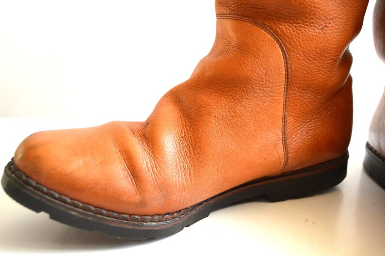 Puede incluir: Una bota de cuero color canela con superficie texturizada, punta redondeada y una suela gruesa de goma negra con tacos. La bota presenta costuras visibles a lo largo del cerco y una costura vertical en el lateral de la ca&ntilde;a, con acabado granulado.