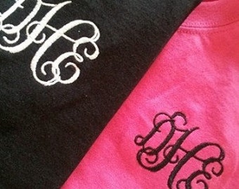 May include: Two t-shirts, one black and one pink, with the same monogram embroidered on each. The monogram is a stylized "DHC" in white on the black shirt and black on the pink shirt.