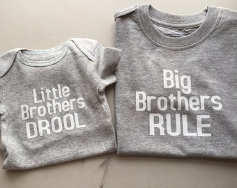 May include: Two gray t-shirts with white text. The smaller shirt says "Little Brothers Drool". The larger shirt says "Big Brothers Rule".