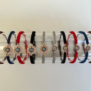 May include: A set of ten colourful string bracelets with a gold evil eye charm. Each bracelet is made with a different colour string, including red, blue, black, and grey.