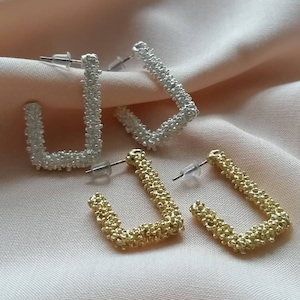 May include: Two pairs of earrings, one pair is gold and the other is silver. Both pairs are made of small, textured beads that form a square shape.