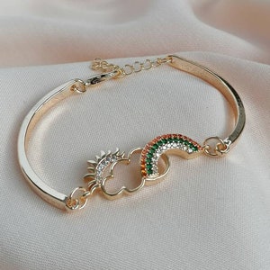 Puede incluir: Una pulsera de oro con un dije que representa un sol, una nube y un arcoíris. El dije está decorado con diamantes de imitación transparentes y verdes.