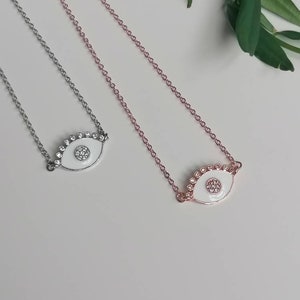 May include: Two delicate necklaces with white enamel evil eye pendants. The pendants are adorned with sparkling clear rhinestones. One necklace has a silver chain and the other has a rose gold chain.