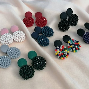 Puede incluir: Surtido de pendientes de cuentas con cuentas de varios colores y respaldo de tela. Los pendientes se muestran sobre una superficie de tela blanca.