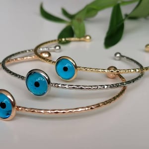 May include: Three gold, silver, and rose gold bangle bracelets with blue evil eye charms. The bracelets are stacked on top of each other.