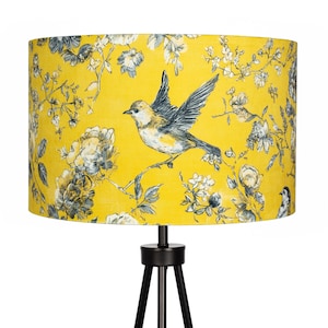 May include: A yellow lampshade with a bird and floral pattern. The lampshade displays a detailed illustration of a bird in flight, surrounded by grey and white flowers and foliage. The lampshade is on a black tripod lamp base.