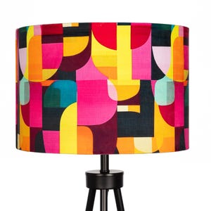May include: A vibrant lampshade with a geometric design in pink, yellow, orange, and teal. The lampshade is cylindrical and sits on a black tripod lamp base. The pattern consists of abstract shapes.