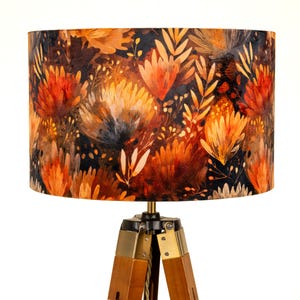 May include: A lampshade featuring a floral design in orange, brown, and black hues. The lampshade is cylindrical and is supported by a wooden tripod base with brass detailing. The botanical pattern includes stylized flowers and foliage.